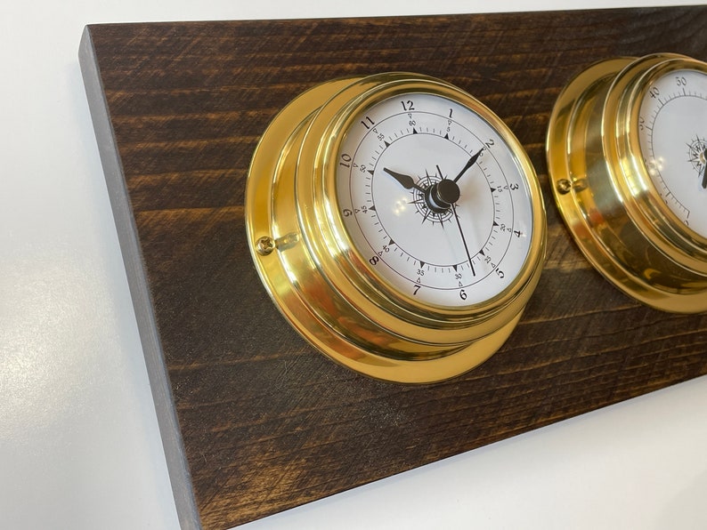 Puede incluir: Dos relojes de barco de lat&oacute;n con caras blancas y manecillas negras montados en una placa de madera marr&oacute;n oscuro.