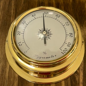Op de afbeelding: Een messing hygrometer met een wit gezicht en zwarte markeringen. De naald wijst naar 50%. De hygrometer is ge&euml;tiketteerd "Hygrometer" en heeft een percentageteken.