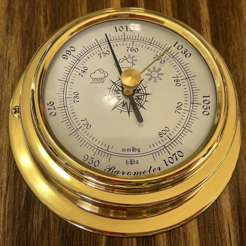 Op de afbeelding: Een messing barometer met een wit gezicht en een zwarte naald. Het gezicht heeft een kompasroos, weersymbolen en markeringen voor millimeters kwik en hectopascal. De tekst "mmHg hPa Barometer" is op het gezicht gedrukt.