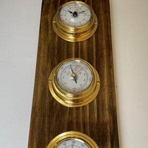 Op de afbeelding: Een houten wandplaat met drie messing omrande meters. De bovenste meter is een klok met zwarte wijzers en Romeinse cijfers. De middelste meter is een barometer met een wit gezicht en zwarte markeringen. De onderste meter is een kompas met een wit gezicht en zwarte markeringen.