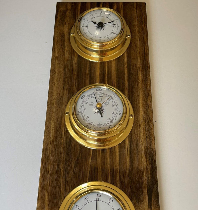 Op de afbeelding: Een houten wandplaat met drie messing omrande meters. De bovenste meter is een klok met zwarte wijzers en Romeinse cijfers. De middelste meter is een barometer met een wit gezicht en zwarte markeringen. De onderste meter is een kompas met een wit gezicht en zwarte markeringen.