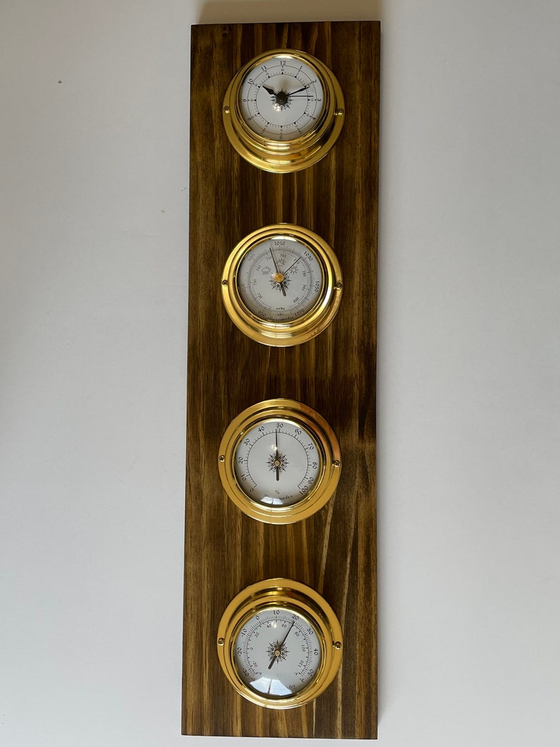 Op de afbeelding: Een set van vier nautische klokken met messing frames gemonteerd op een donker houten paneel. De klokken omvatten een klok, een barometer, een thermometer en een hygrometer.