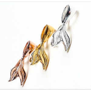 Puede incluir: Tres colgantes de cola de sirena de metal en oro rosa, dorado y plateado. Los colgantes están dispuestos en diagonal sobre un fondo blanco. Cada colgante tiene un lazo en la parte superior para sujetarlo a la joyería.