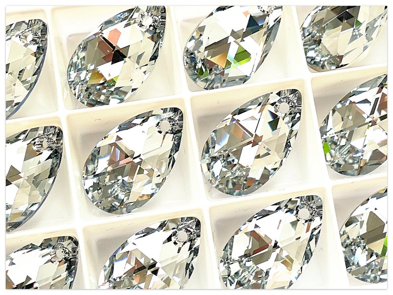 Puede incluir: Primer plano de m&uacute;ltiples cristales transparentes facetados en forma de l&aacute;grima. Cada cristal est&aacute; colocado en un compartimento blanco. Los cristales tienen un agujero en la parte superior para ensartar o sujetar. Los cristales reflejan la luz, creando un efecto brillante.