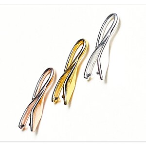 Könnte beinhalten: Drei Metall-Schmuckzubehörteile in Roségold, Gold und Silber. Jedes Zubehörteil hat ein gebogenes, bandartiges Design mit einer kleinen Schlaufe am unteren Ende. Die Zubehörteile sind vor einem weißen Hintergrund platziert.