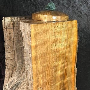 Op de afbeelding: Een handgemaakte houten doos met een deksel. De doos is gemaakt van lichtbruin hout met een natuurlijke, getextureerde schors aan één kant en een glad, gepolijst oppervlak aan de andere kant. Het deksel is bekroond met een kleine, groene steen.