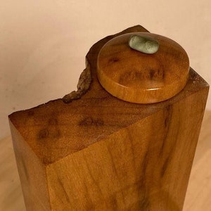 May include: A wooden box with a lid, the lid is made of wood and has a green stone on top. The box is sitting on a wooden surface.