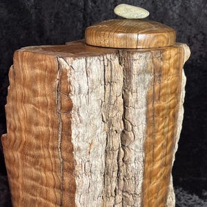 May include: A wooden box with a natural, textured finish. The box has a lid with a small stone on top. The wood grain is visible, showcasing shades of brown and tan. The box is a unique decorative item.