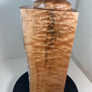 以下が含まれることがあります： 天然の木目が美しい木製の骨壺。 骨壺には蓋があり、上には小さな灰色の石が乗っています。 木目は、濃い茶色の縞模様のある波状のパターンです。 骨壺は長方形で、わずかに湾曲した側面があります。