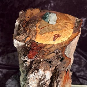 Może przedstawiać: Ręcznie wykonane drewniane pudełko z pokrywką, o naturalnym, rustykalnym designie. Pudełko wykonane jest z różnych rodzajów drewna, prezentując mieszankę jasnych i ciemnych brązowych odcieni. Mały, szaro-zielony kamień jest przymocowany do pokrywki. Pudełko ma około 15 cm wysokości.