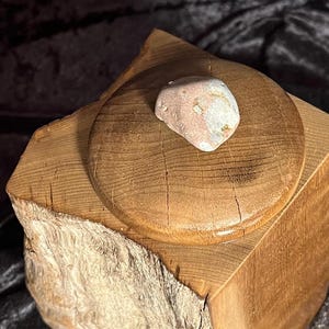 May include: A wooden box with a lid, made from a tree trunk. The lid has a round, smooth surface and a pink and white stone is placed on top of it.