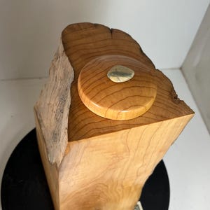 May include: A handcrafted wooden box with a natural, light brown finish. The box features a rounded lid with a small, embedded stone. The wood grain is visible, and the box has a rustic, organic aesthetic. The box is approximately 6 inches tall.