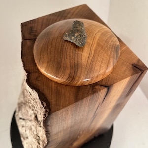 May include: A wooden box with a lid, made from a tree trunk with a natural bark edge. The lid is round and has a small, dark grey stone on top.