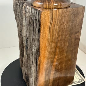 Puede incluir: Una caja de madera hecha a mano con un borde natural en un lado y un acabado liso y pulido en el otro. La tapa está rematada con una pequeña piedra gris. La caja se muestra sobre una superficie negra.