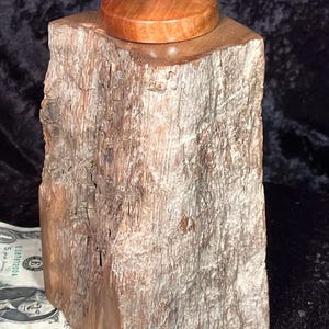 May include: A rustic wooden box with a natural, textured finish. The box is topped with a circular wooden lid and a small, oval-shaped stone. The wood has a light brown colour with variations in tone.