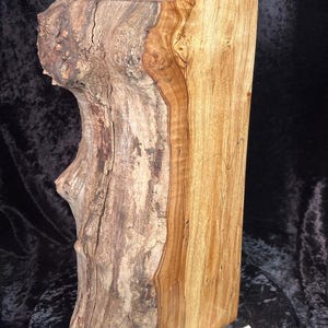 Puede incluir: Urna de madera artesanal con una estética rústica natural. La urna presenta un cuerpo rectangular con un acabado liso marrón claro en un lado y una superficie texturizada similar a la corteza en el otro. Una tapa redonda con una pequeña piedra en la parte superior completa el diseño.