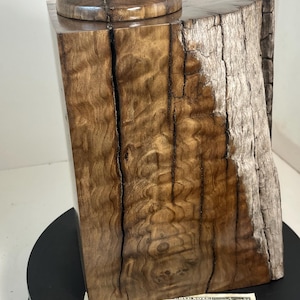May include: A handcrafted wooden box with a lid, featuring a natural wood grain pattern. The box is rectangular with a rounded lid, topped with a small stone. The wood has a rich, warm brown color. The box is displayed on a black surface.