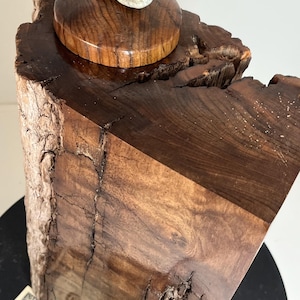 May include: A handcrafted wooden sculpture with a natural, rustic aesthetic. The sculpture features a dark brown, polished wooden base with a circular, polished wooden top. A small, light-colored stone rests on the top, adding a natural contrast to the wood.