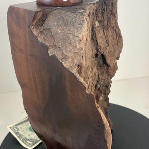 Può includere: Una scatola di legno fatta a mano con finitura marrone scuro e un bordo naturale e grezzo. Il coperchio è un pezzo di legno lucido e circolare, sormontato da una piccola pietra liscia. La scatola è un oggetto decorativo unico.