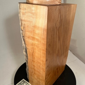 以下が含まれることがあります： 自然な縁のある木材の一枚板で作られた、蓋付きの木箱。蓋の上には小さな薄い灰色の石が置かれている。箱は黒い表面の上に置かれている。