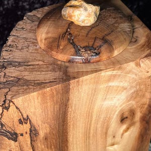 May include: A wooden box with a lid, made from a single piece of wood with a natural wood grain pattern. The lid has a round shape and a small, light brown stone on top.