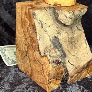 May include: A handcrafted wooden box with a lid, featuring a natural, rustic design. The box is made from light and dark brown wood with unique grain patterns and a small, light green stone on top of the lid. The box is a decorative item.