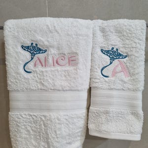 May include: Two white bath towels with blue manta ray embroidery. One towel has the name "Alice" embroidered in pink.