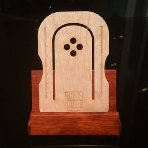 Puede incluir: Un soporte de teléfono de madera con un recorte para un teléfono y una regla a cada lado. El soporte tiene un logotipo que dice "SUMI WINE" y "Hecho en Tailandia".