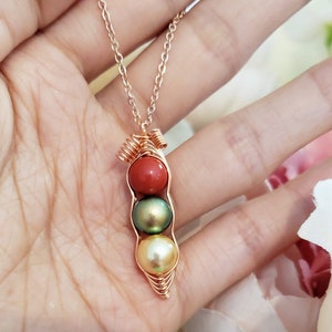 May include: A gold necklace with a pea pod pendant featuring three pearls. The top pearl is red, the middle pearl is green, and the bottom pearl is white.