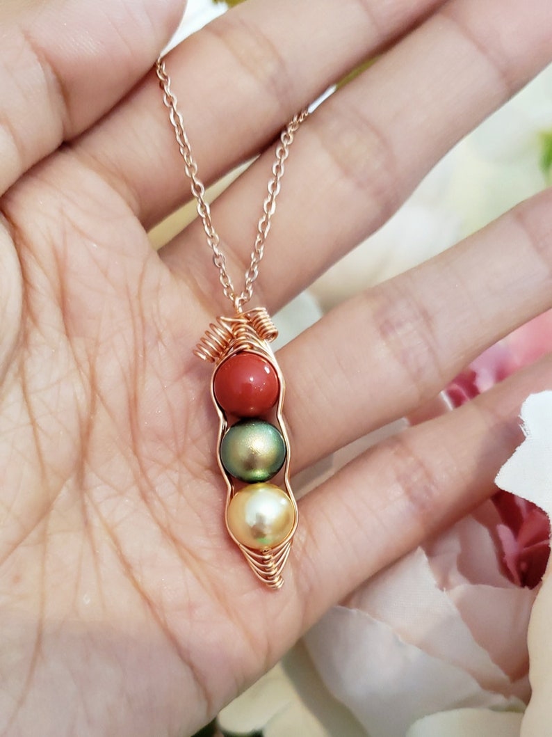 May include: A gold necklace with a pea pod pendant featuring three pearls. The top pearl is red, the middle pearl is green, and the bottom pearl is white.