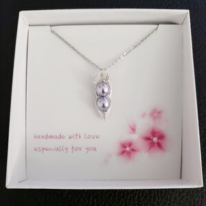 May include: A silver necklace with a pea pod pendant featuring two purple pearls. The necklace is in a white box with a pink floral design and the text "handmade with love especially for you".