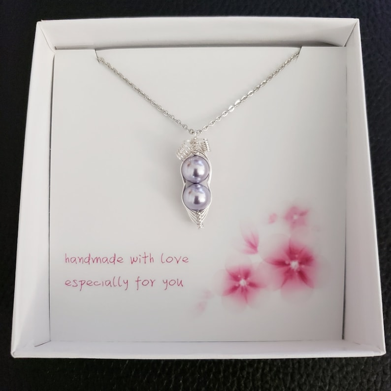 May include: A silver necklace with a pea pod pendant featuring two purple pearls. The necklace is in a white box with a pink floral design and the text "handmade with love especially for you".