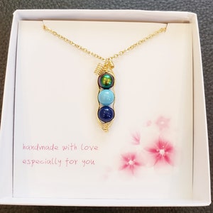 May include: A gold necklace with a pea pod charm featuring three blue and green beads. The necklace is in a white box with the text "handmade with love especially for you".