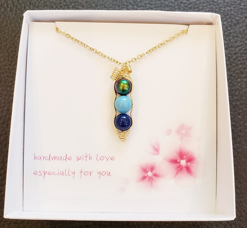 May include: A gold necklace with a pea pod charm featuring three blue and green beads. The necklace is in a white box with the text "handmade with love especially for you".