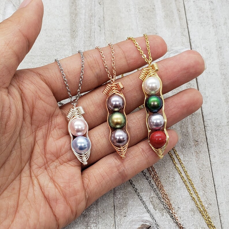 May include: Three necklaces with pea pod pendants featuring different colored pearls. The pendants are made of wire and feature a silver, green, and red pearl.