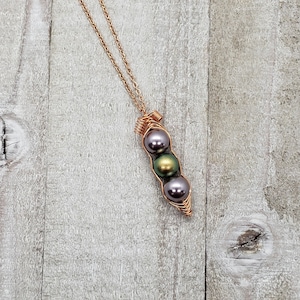 May include: A copper wire necklace with a pea pod charm. The charm has three pearls inside: one dark green, one iridescent green, and one dark purple.