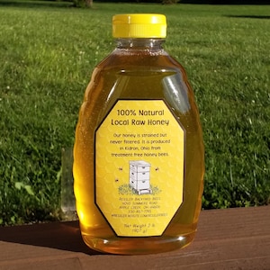 May include: A clear plastic bottle of 100% natural local raw honey. The label is yellow and black with the text "100% Natural Local Raw Honey" and information about the honey and the bee farm. The bottle is sitting on a wooden surface.