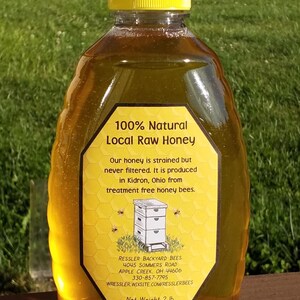 May include: A clear plastic bottle of 100% natural local raw honey. The bottle has a yellow lid and a yellow label with a beehive illustration and text that reads "100% Natural Local Raw Honey", "Our honey is strained but never filtered. It is produced in Kidron, Ohio from treatment free honey bees.", "RESSLER BACKYARD BEES 4045 SOMMERS ROAD APPLE CREEK, OH 44606 330-857-7795 WRESSLER. WIXSITE.COM/RESSLERBEES", and "Net Weight 2 lb (907 g)".