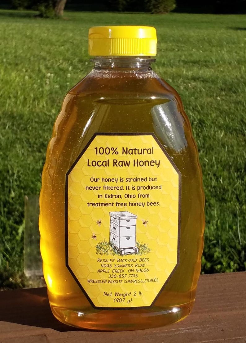 May include: A clear plastic bottle of 100% natural local raw honey. The bottle has a yellow lid and a yellow label with a beehive illustration and text that reads "100% Natural Local Raw Honey", "Our honey is strained but never filtered. It is produced in Kidron, Ohio from treatment free honey bees.", "RESSLER BACKYARD BEES 4045 SOMMERS ROAD APPLE CREEK, OH 44606 330-857-7795 WRESSLER. WIXSITE.COM/RESSLERBEES", and "Net Weight 2 lb (907 g)".