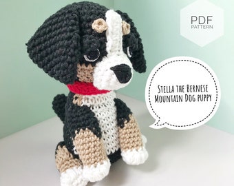 Może przedstawiać: Pluszowa zabawka amigurumi szczeniaka bernenskiego psa górskiego wykonana na szydełku, z czarnym, białym i brązowym futrem, w czerwonym kolarzu. Zabawka siedzi na białym tle. Tekst "Stella the Bernese Mountain Dog Puppy" znajduje się w dymku.