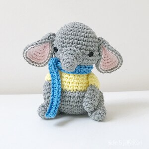 May include: A gray crocheted elephant wearing a blue and yellow striped scarf. The elephant has pink ears and black eyes.