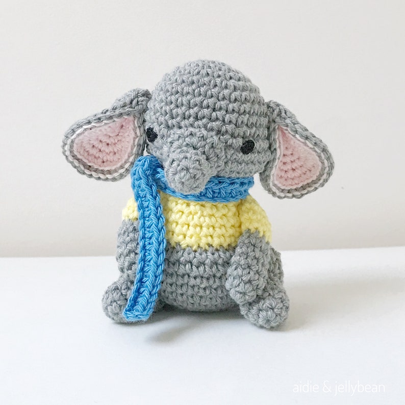May include: A gray crocheted elephant wearing a blue and yellow striped scarf. The elephant has pink ears and black eyes.