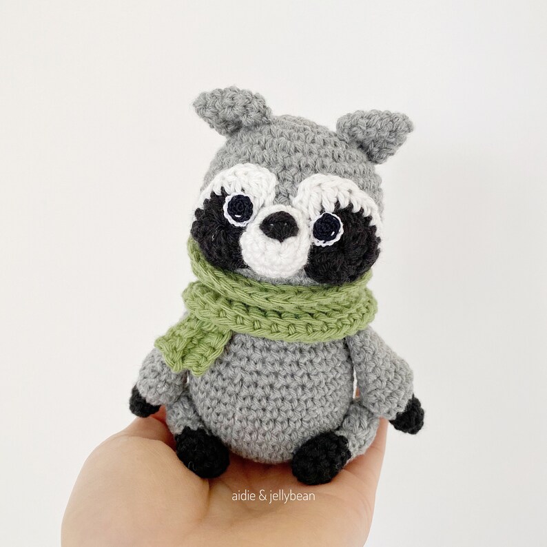 May include: A crocheted raccoon wearing a green scarf. The raccoon has black eyes, a white nose, and black and white markings around its eyes.