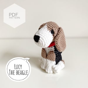 Può includere: Giocattolo amigurumi a uncinetto a forma di beagle di nome Lucy. Il cane è marrone, bianco e nero con un collare rosso. Il giocattolo è seduto su una superficie bianca. Il testo "Lucy The Beagle" è in una nuvola di dialogo.