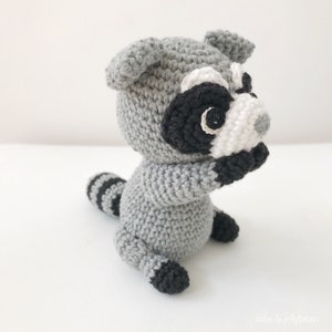 May include: A crocheted raccoon plush toy with gray fur, black eyes, a white nose, and a black and white striped tail.