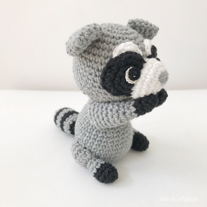 May include: A crocheted raccoon plush toy with gray fur, black eyes, a white nose, and a black and white striped tail.