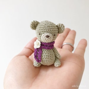 May include: A small crocheted teddy bear wearing a purple scarf with a white crocheted flower.