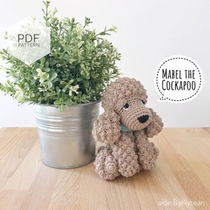 Puede incluir: Un perro Cockapoo de crochet marr&oacute;n y blanco llamado Mabel. El perro est&aacute; sentado en una superficie de madera con una planta verde en una maceta plateada detr&aacute;s de &eacute;l. El texto "Mabel the Cockapoo" est&aacute; en un bocadillo de di&aacute;logo sobre el perro.