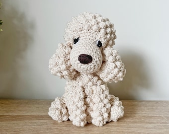May include: A handmade, beige crochet poodle dog. The dog has a textured, curly appearance, with a dark brown nose and black eyes. The poodle is sitting upright, with its front paws together. The background is a light-colored wall and a wooden surface.