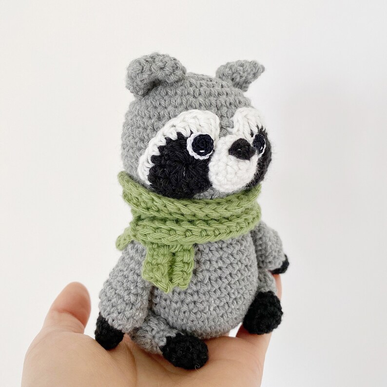May include: A gray crocheted raccoon wearing a green scarf. The raccoon has black eyes and a white nose.